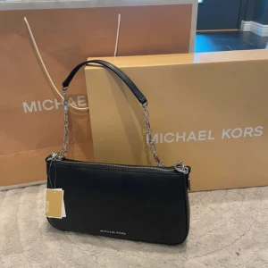 Svart handväska från Michael Kors - Stilren svart handväska från Michael Kors i slät skinn med silverfärgade detaljer. Väskan har en kedjerem med MK-logga, dragkedja upptill och minimalistisk design. Perfekt storlek för det viktigaste och ger en lyxig känsla till din outfit.