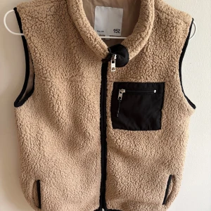 Beige fleeceväst från 157 - Mysig beige fleeceväst från 157 med svart bröstficka och dragkedja framtill. Västen har hög krage och svarta detaljer vid fickor och kanter. Perfekt för lager-på-lager och chill dagar.