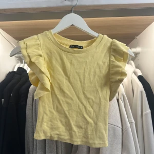 Gul croppad topp med volangärmar Zara - Superfin ljusgul croppad topp från Zara med volangdetaljer på axlarna. Toppen är ribbad och har en rund halsringning. Perfekt för dig som vill ha en söt och trendig look till sommaren.