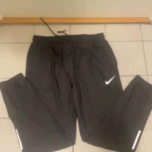 Svarta träningsbyxor från Nike - Svarta träningsbyxor från Nike med vitt logotryck på låret och reflexdetaljer vid bensluten. Byxorna har elastisk midja med snörning och är tillverkade i ett lätt syntetmaterial som passar perfekt för träning.