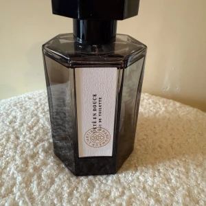 L'Artisan Parfumeur Thé en Douce EdT - Snygg parfymflaska i mörkgrått glas med svart, åttakantigt lock. Märkt med L'Artisan Parfumeur och doften Thé en Douce. Flaskan har en elegant, modern design och vit etikett framtill med logotyp. Perfekt för dig som gillar unika och stilrena dofter.