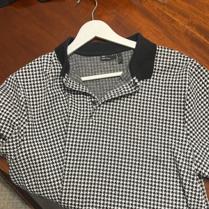 Svartvit rutig pikétröja från ASOS  - Snygg pikétröja från ASOS DESIGN i storlek XL med klassiskt svartvitt hundtandsmönster. Tröjan har svart krage, korta ärmar och knäppning framtill. Perfekt för dig som gillar en stilren och trendig look. Hör av vid frågor!🙌🏽 Alltid öppen till prisförslag!