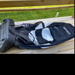 Svart och grå resväska från Open - Stor och rymlig golfbag från Open i svart och grått med hård botten och flera fack med dragkedja. Perfekt för att packa sportutrustning eller större saker. Praktiska handtag och robust design för enkel transport.