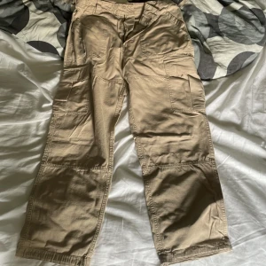 Beiga cargopants med fickor, YOKE. - Säljer ett par beiga cargopants från Blåkläder med flera stora fickor på sidorna och bak. Byxorna har en loose passform och är tillverkade i slitstark bomull. Perfekta för dig som gillar praktiska och rymliga byxor med streetkänsla. Från Yoke.