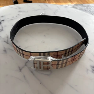 Burberry bälte - Snyggt Burberry-bälte med klassiskt beige, brunt och rött rutmönster. Bältet har ett stort silvrigt B-spänne och insidan är svart med präglade detaljer. Perfekt accessoar för att lyfta din outfit med en lyxig touch.