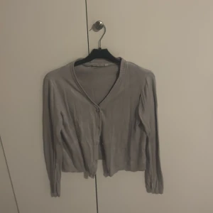 Grå kofta från KappAhl med knappar - En basic grå kofta från KappAhl med v-ringning och knäppning framtill. Långärmad och i mjukt, ribbat material som känns skönt mot huden. Perfekt att slänga över en t-shirt eller linne för en chill look.
