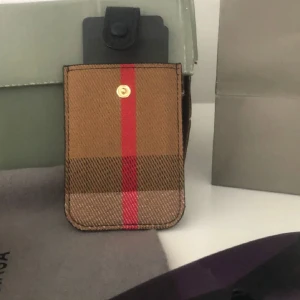 Rutig plånbok från Burberry - Snygg liten plånbok från Burberry i klassiskt rutigt mönster med bruna och beige toner samt en röd rand. Plånboken har svart knappstängning och är tillverkad i tåligt material. Perfekt för kort och småsaker, stilren och ikonisk design.