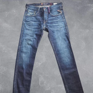 Replay jeans - Snygga blå Replay Anbass jeans i samarbete med FC Barcelona. Klassisk femficksmodell med raka ben, coola slitningar och detaljer i rött och blått. Tillverkade i Italien av jeansmaterial med kontrastsömmar och patch bak i midjan.
