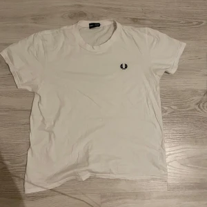 Vit Fred Perry t-shirt i bomull - Vit t-shirt från Fred Perry med klassisk svart broderad lagerkrans på bröstet. Enkel och stilren design med rund hals och korta ärmar. Tillverkad i mjuk bomull som känns skön mot huden. Perfekt för en clean och avslappnad look.