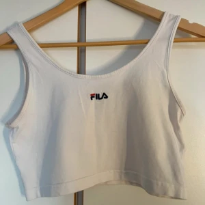 Vit croppad linnetopp från FILA - Snygg vit croppad linnetopp från FILA med broderad logga framtill. Toppen har breda axelband och ärmlös design, perfekt för en sportig och avslappnad stil. Tillverkad i mjuk bomull som känns skön mot huden.