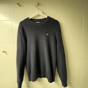 Mörkblå stickad tröja med brodyr - Mörkblå stickad tröja med rund hals och diskret broderad detalj på bröstet. Tröjan har lång ärm och ribbade muddar vid ärmslut och nederkant. Perfekt för lager-på-lager och enkel att matcha med jeans eller chinos.