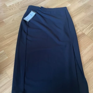 Svart långkjol från New Yorker - Stilren svart långkjol från New Yorker med rak passform och diskret slits på sidan. Kjolen är enkel och clean, perfekt om du gillar minimalistisk stil. Tillverkad i ett mjukt syntetmaterial som känns skönt mot huden.