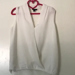 Vit omlottopp med spetsrygg - Säljer en vit ärmlös omlottopp  med djup v-ringning och elastisk nederkant. Baksidan har en snygg spetsdetalj i triangelform som ger toppen en extra touch. Perfekt för dig som gillar stilrena och feminina plagg.