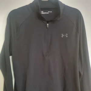 Svart tröja med dragkedja från Under Armour - Svart tröja från Under Armour med halv dragkedja och långärmad design. Perfekt för träning eller vardagsbruk. Tröjan har en diskret logga på bröstet och i nacken.