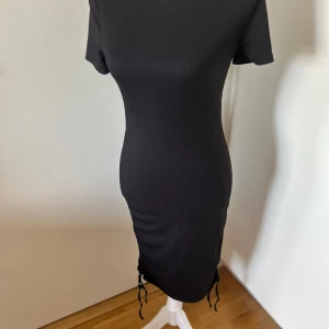 Svart ribbad klänning - Snygg svart ribbad klänning med korta ärmar och åtsittande passform. Klänningen har en knytning på sidan som ger en fin detalj och form. Perfekt för en stilren look.