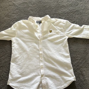 Vit skjorta från Lyle & Scott Junior - Säljer en stilren vit skjorta från Lyle & Scott Junior. Skjortan har en klassisk krage och knappar framtill. Den är dekorerad med en broderad logotyp i gult på bröstet. Perfekt för en snygg och avslappnad look.