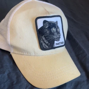 Beige keps med pantermärke - Snygg beige keps med ett broderat märke av en panter på framsidan. Kepsen har en klassisk design med justerbar passform och vit mesh på baksidan för extra ventilation.