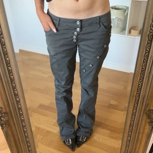 Bootcutjeans med knappar - Snygga grå byxor med unika knappdetaljer längs sidorna och framtill. Byxorna har en bootcut-stil och är lågmidjade, vilket ger en cool och avslappnad look. Perfekta för dig som vill ha något annorlunda i garderoben. Midjemått 40cm