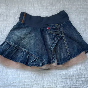 Jeanskjol med volang och tyll - Snygg jeanskjol med volangdetaljer och tyllkant. Kjolen har en elastisk midja i mörkare tyg och en omlott-design framtill. Perfekt för en trendig look!