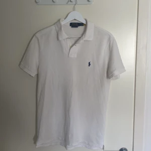 Vit pikétröja från Ralph Lauren - Säljer en klassisk vit pikétröja från Ralph Lauren i custom slim fit. Tröjan har korta ärmar och den ikoniska broderade logotypen i mörkblått på bröstet. Perfekt för en stilren och avslappnad look.