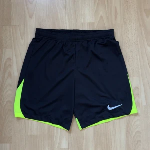 Svarta träningsshorts från Nike - Snygga svarta träningsshorts från Nike med neongula detaljer på sidorna. Perfekta för sport och träning. De har en elastisk midja för bekväm passform och är tillverkade i ett lätt och andningsbart material.