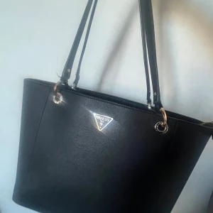 Svart handväska från Guess - Snygg svart handväska från Guess med stilren design. Väskan har dragkedja på baksidan och rymligt inre med flera fack. Perfekt för att bära med sig allt man behöver under dagen. Skriv för fler frågor eller mått💕Den är köpt från Best Sexret, orginalpris: 1200kr