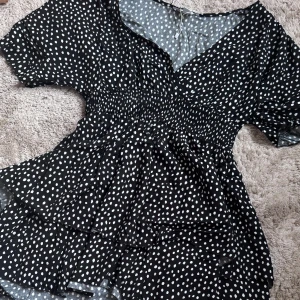 Svart prickig klänning från Amisu  - Säljer en kort svart klänning med vita prickar från Amisu. Klänningen har en v-ringad design och korta ärmar. Den har en smickrande passform med volanger som ger en lekfull touch. Oanvänd