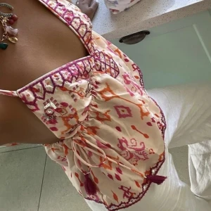 Mönstrad ärmlös topp med broderi - Säljer en färgglad ärmlös topp med v-ringning och vackert broderi. Toppen har ett livfullt mönster i rosa, orange och beige med justerbara axelband och knytning framtill. Perfekt för sommardagar!