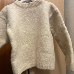 Beige/krämvit stickad tröja från Dobber - Mysig beige stickad tröja från Dobber. Perfekt för kyliga dagar med sin mjuka och varma känsla. Tröjan har en klassisk design med rund hals och långa ärmar.