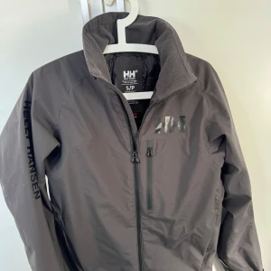 Grå jacka från Helly Hansen - Säljer en grå jacka från Helly Hansen med dragkedja och hög krage. Jackan har en diskret logga på bröstet och är tillverkad i ett syntetmaterial. Perfekt för kyligare dagar.