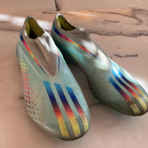 Flerfärgade fotbollsskor - Säljer ett par snygga fotbollsskor i flerfärgat mönster med dominanta blå och silver detaljer. Skorna har en strömlinjeformad design utan snörning för en tight passform. Perfekta för den som vill sticka ut på planen! Sjukt sköna skor