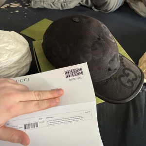 Svart keps från Gucci - Snygg svart keps från Gucci med diskret mönster av märkets logotyp. Kepsen har en klassisk design med böjd skärm och justerbar rem baktill för perfekt passform. Perfekt för att ge en lyxig touch till din outfit. Nypris 5400