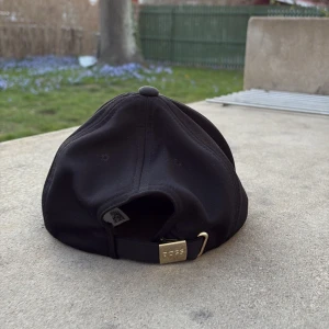 Svart keps från BOSS - Snygg svart keps från BOSS med diskret mönster och justerbart spänne i guld. Perfekt för en stilren look.