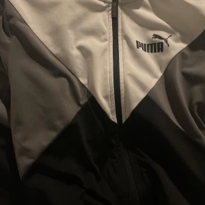 Svart och vit vindjacka från Puma - Säljer en stilren svart och vit vindjacka från Puma med dragkedja framtill. Jackan har en sportig look med Pumas logga på bröstet. Perfekt för en aktiv livsstil.