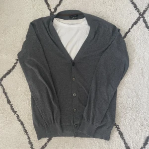 Grå kofta från Massimo Dutti - Säljer en elegant grå kofta från Massimo Dutti, tillverkad i en lyxig blandning av bomull, silke och kashmir. Koftan har knappar framtill och ribbade muddar vid ärmslut och nederkant. Perfekt för en stilren look.