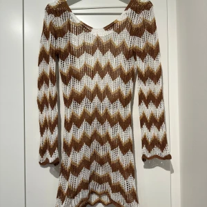 Brun och vit stickad klänning - Säljer en snygg stickad klänning i brunt och vitt med ett zigzag-mönster. Klänningen har långa ärmar och en knytning i ryggen. Perfekt för en trendig look!