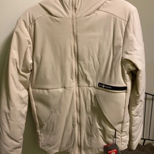 Beige jacka från sail Racing  - Säljer en stilren beige jacka från sali Racing. Jackan har en dragkedja framtill och en praktisk huva. Perfekt för kyligare dagar med sin långa ärmar och bekväma passform.