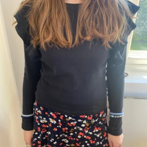 Svart ribbad tröja från SRNLT - Snygg svart ribbad tröja från SRNLT med volangdetaljer på axlarna. Perfekt för att ge en stilren look med en twist. Tröjan har långa ärmar och en bekväm passform. Ett måste i garderoben för den som gillar att mixa och matcha! perfekt till jeans. Super trendig just nu. Passar även till kjolen min modell har på sig som jag också säljer❤️🌸😻. Liten i storlek.  Den är ogentligen i barn storlek 156-164