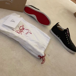 Svarta skor från Christian Louboutin (med kvittot) - Snygga svarta sneakers från Christian Louboutin med ikoniska röda sulor och silverfärgade nitar. Skorna har en rund tå och snörning framtill. Perfekta för att ge din stil en edgy touch.