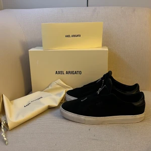 Svarta sneakers från Axel Arigato - Snygga svarta sneakers från Axel Arigato med dragkedja på ovansidan. Skorna har en vit sula som ger en stilren kontrast. Perfekta för en trendig look.