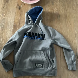 Grå hoodie från Hoodrich - Säljer en grå hoodie från Hoodrich med blått foder i huvan och svart text på framsidan. Den har en stor ficka framtill och en etikett med texten 'From Nothing to Something'. Perfekt för streetwear-stilen.