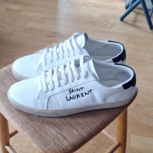 Vita sneakers från Saint Laurent - Snygga vita sneakers från Saint Laurent med svart detalj vid hälen. Skorna har snörning och en rund tå. Perfekta för en stilren look. Dom är lite creased men annat än det så är dom i princip nyskick. Boxen är ganska skadad men kan skickas med om önskas. Pris ej hugget i sten. Skriv vid funderingar🙌