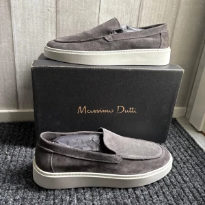 Gråa mockaloafers från Massimo Dutti - Snygga gråa loafers i mocka från Massimo Dutti. Skorna har en stilren design med en vit sula som ger en modern touch. Perfekta för en avslappnad men ändå elegant look. Aldrig använda endast testade, köpta i Göteborg och var för stora