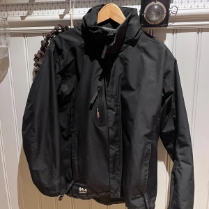 Svart vindjacka från Helly Hansen - Säljer en stilren svart vindjacka från Helly Hansen. Jackan har en dragkedja framtill och en praktisk bröstficka med dragkedja. Perfekt för blåsiga dagar och har en normal passform. Modellen är runt 180 och väger 75kg. 