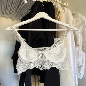 Vit spetslinne i storlek M - Säljer en vacker vit spetslinne med fina detaljer och en liten rosett framtill. Perfekt för att lägga till en romantisk touch till din outfit. Den har en delikat spetsdesign och justerbara axelband.