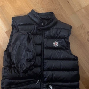 Moncler väst - Säljer en snygg svart dunväst från Moncler med dragkedja. Västen har ett stilrent emblem på bröstet och praktiska fickor med dragkedja. Perfekt för kyliga dagar när du vill hålla stilen.strl S hör av er vid frågor 