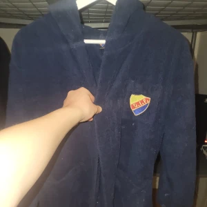 Blå morgonrock med Djurgården-logga - Säljer en blå morgonrock med Djurgården-logga på bröstet och texten 'Djurgården' på ryggen. Rocken har en huva och knyts med band i midjan. Den användes nästan aldrig från mig då jag inte har särskilt mycket sympatier med laget, men för en djurgårdare är det ett drömkap! Den står i 152 men är betydligt större än så, passar ungefär stor 158-164-liten S.