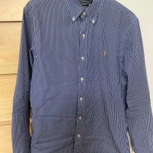 Blå och vit randig skjorta från Polo Ralph Lauren - Snygg blå och vit randig skjorta från Polo Ralph Lauren i slim fit. Skjortan har långa ärmar och knappar framtill samt en broderad logga i gult på bröstet. Perfekt för en stilren look! Inga defekter på skjortan och tveka inte på att höra av er om ni har några frågor:)