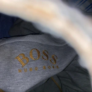 Grå hoodie från Hugo Boss - Säljer en stilren grå hoodie från Hugo Boss med gulddetaljer. Tröjan har en dragkedja framtill och en justerbar huva. Perfekt för en avslappnad look. Kan gå ner i snabbpris 