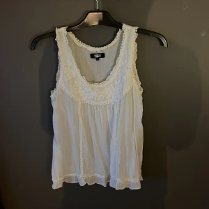 Vit ärmlös blus från Bik Bok - Säljer en söt vit ärmlös blus från Bik Bok med fina broderade detaljer och volangkant. Perfekt för varma dagar eller som ett lager under en kavaj. Blusen har en luftig passform och är gjord i ett lätt material.